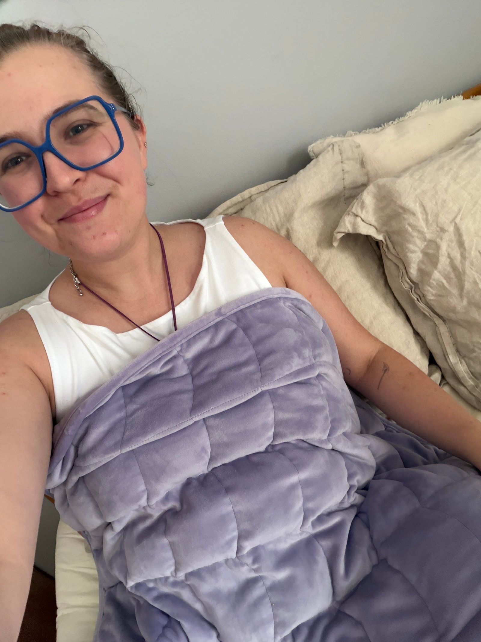 Person with blue glasses and a white top sits on a bed, smiling, covered with a light purple weighted blanket.