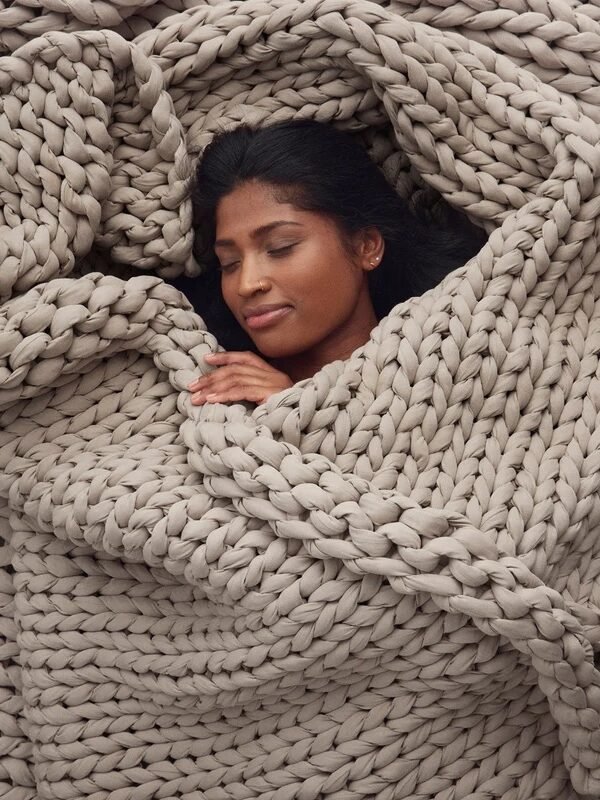 A person lies with eyes closed, wrapped in a large chunky knit beige blanket, appearing comfortable and relaxed.