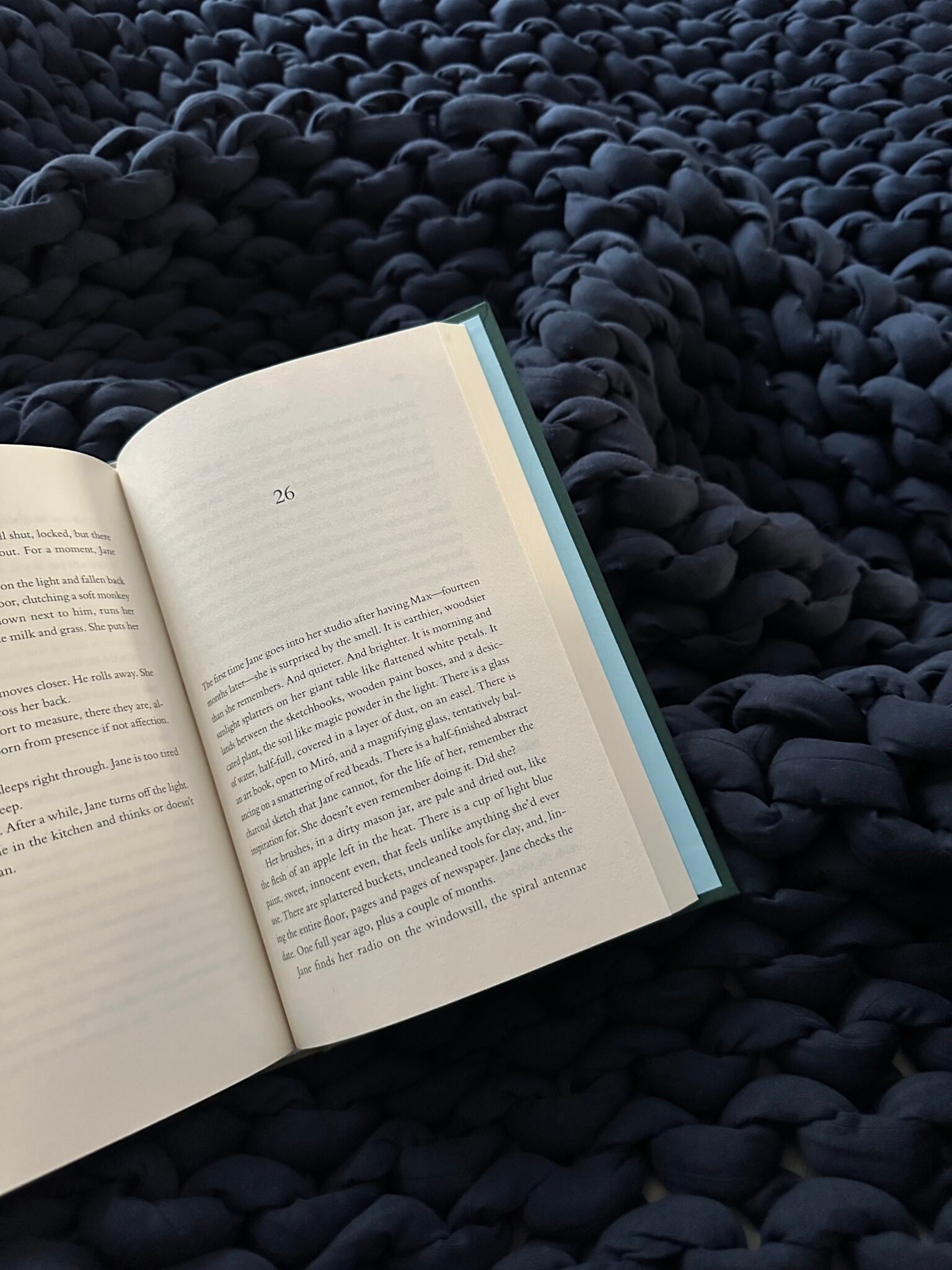 An open book lies on a chunky dark blue knit blanket, displaying page 26 with text visible, in soft natural lighting.