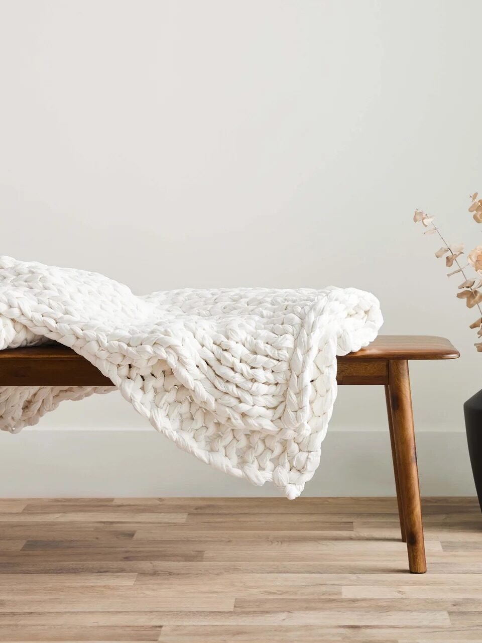 A chunky white knit blanket draped over a wooden bench next to a black vase with dried branches, on a light wood floor against a plain white wall.