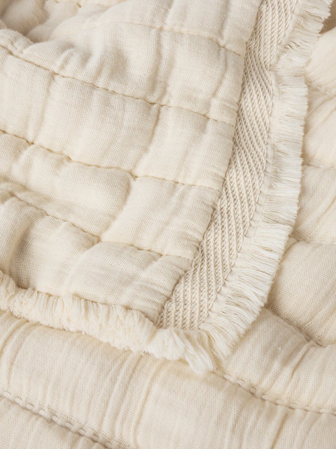 Close-up of a cream-colored, quilted fabric blanket with a textured surface and fringed edge detailing.