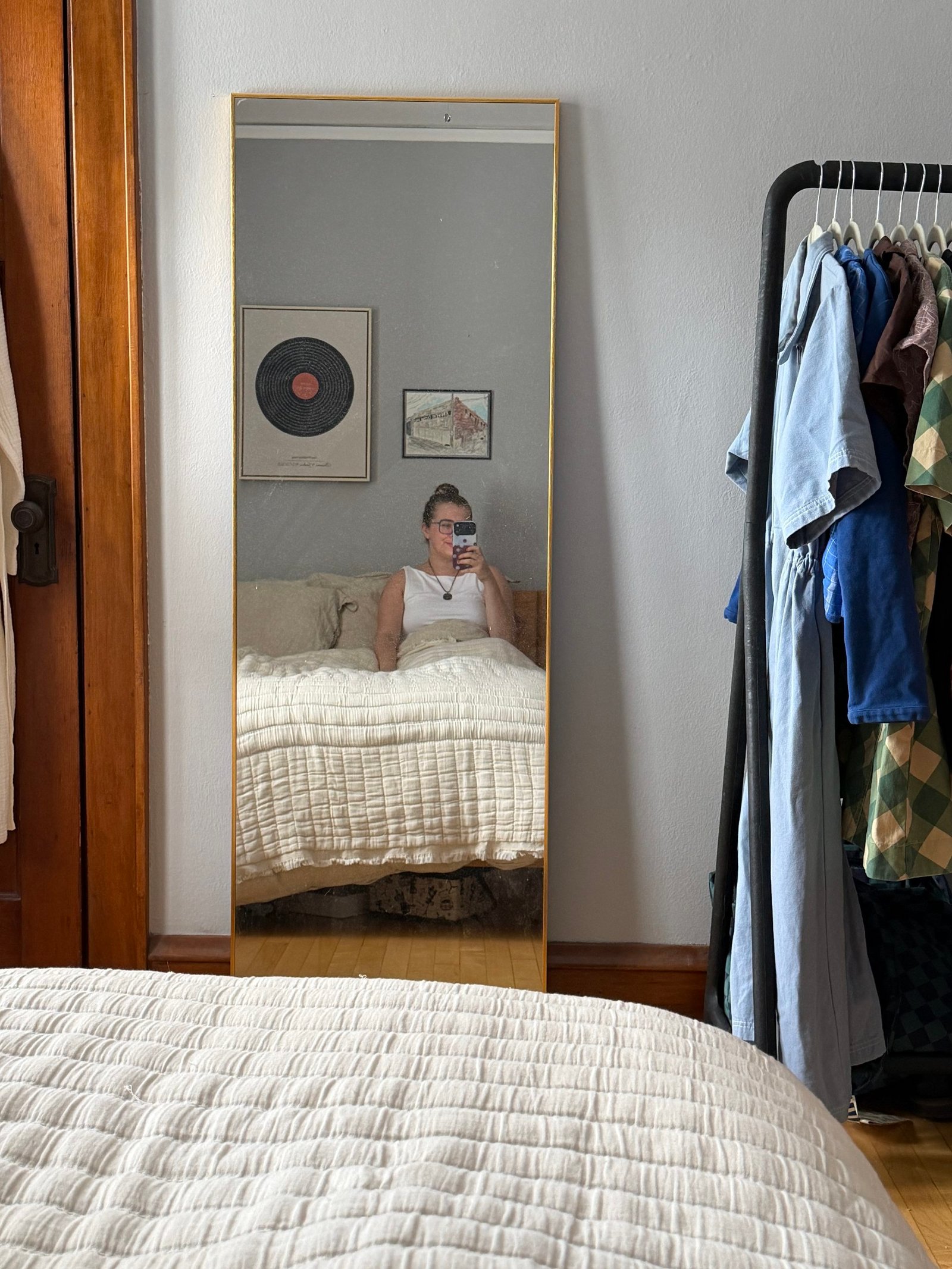 Person sits on a bed taking a mirror selfie in a bedroom with framed art on the wall and a clothing rack to the right.