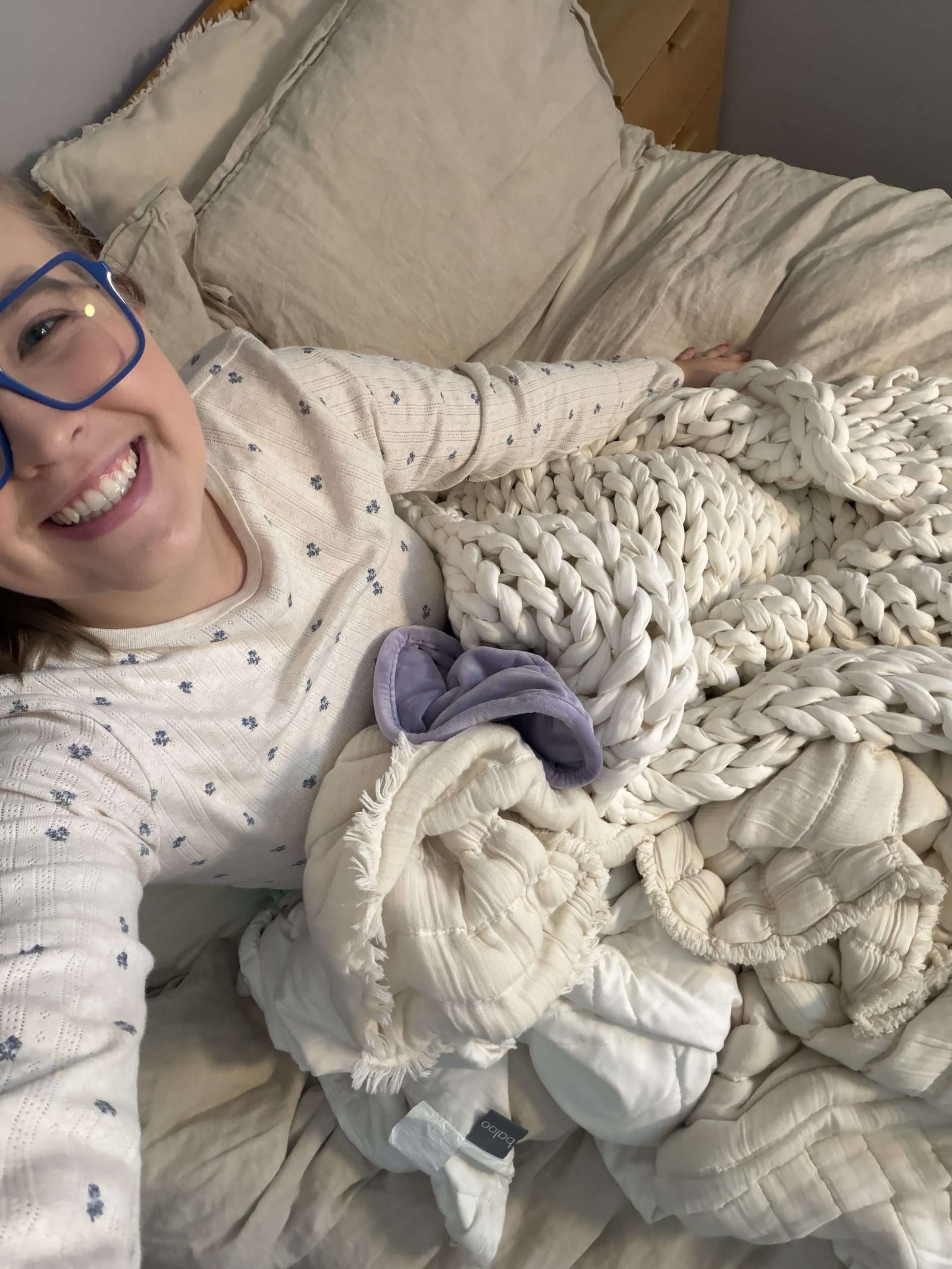 Person wearing glasses and a patterned sweater smiles while lying on a bed with cream-colored blankets and a chunky knit throw.