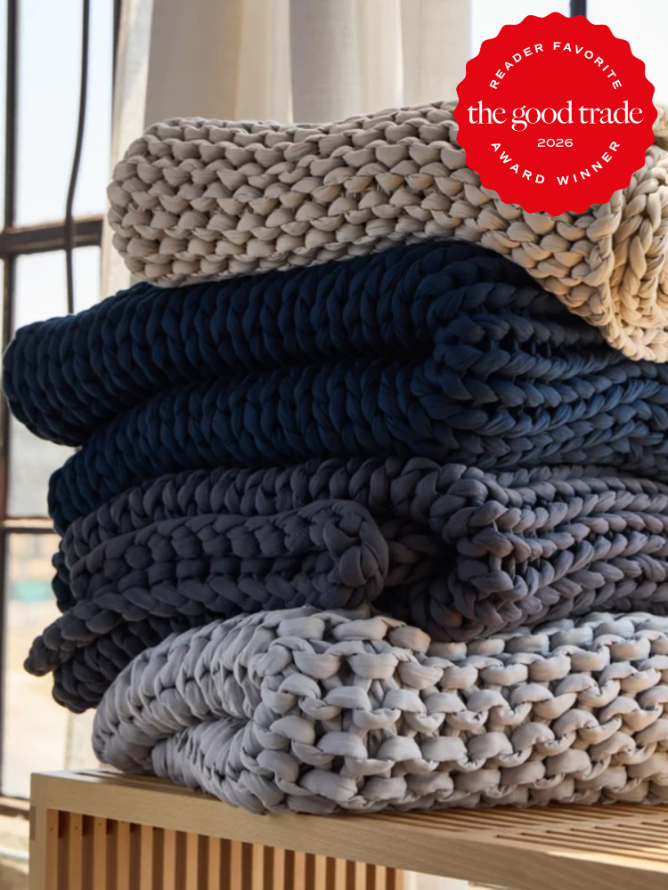 Three chunky knit blankets in cream, navy, and gray are neatly stacked on a wooden surface, with a red “The Good Trade 2026 Award Winner” badge in the top right corner.