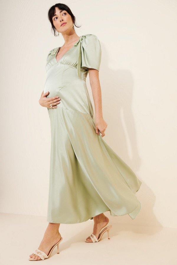 A pregnant woman wearing a light green satin midi dress with short flutter sleeves and heeled sandals.