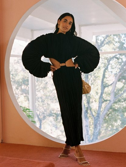 A person wearing a black pleated maxi dress with balloon sleeves and a cut-out waist detail stands in front of a circular window.