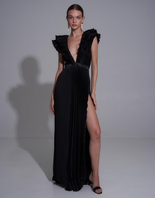 A model in a black, floor-length, pleated gown with a plunging neckline, ruffled shoulders, and a high thigh slit.