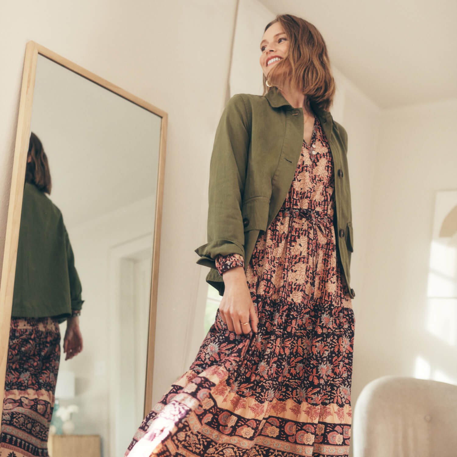 A woman wearing a floral maxi dress and an olive green jacket, reflected in a full-length mirror.