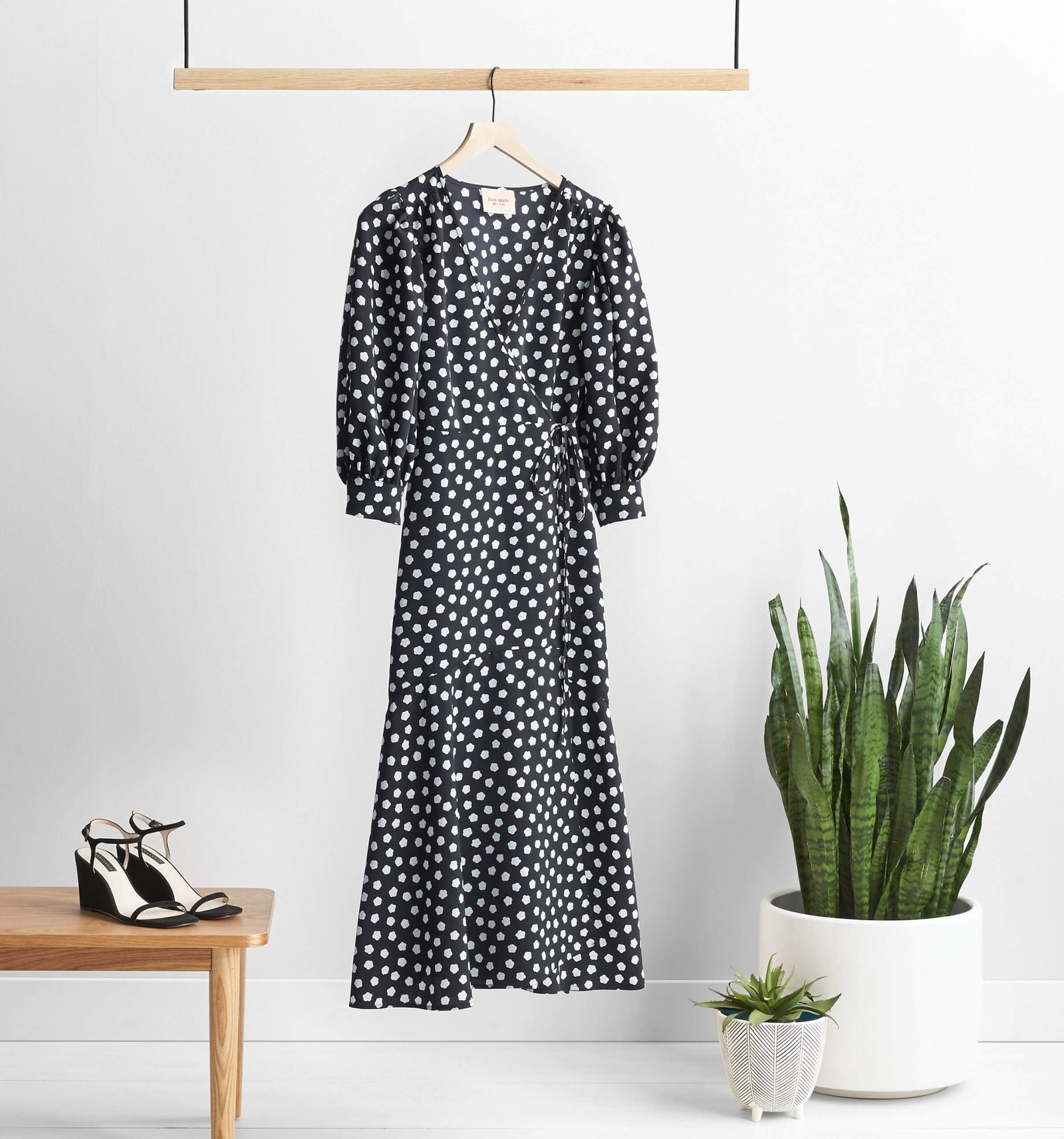 Black midi wrap dress with a white floral pattern hanging on a wooden rack, with black wedge sandals on a side table.