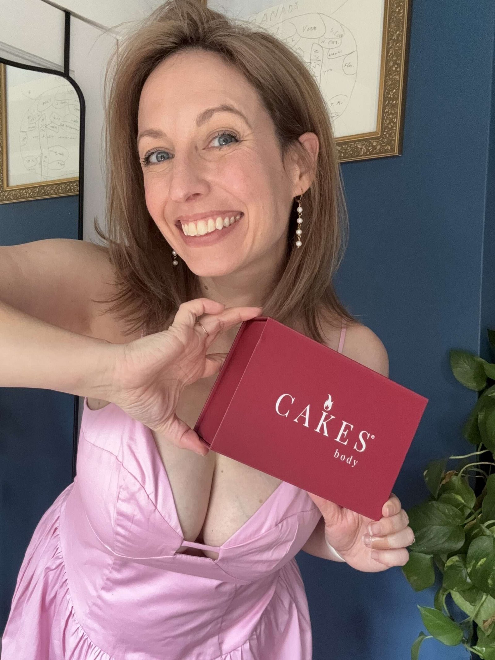 Woman in a pink dress smiles and holds a red box labeled "CAKES body" in front of a blue wall with framed diagrams and a green plant.