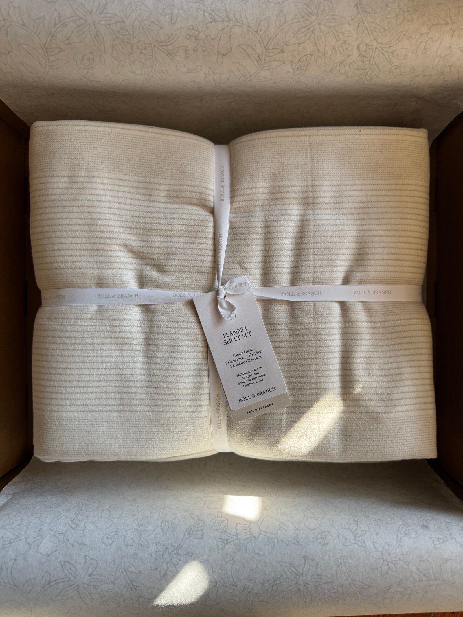 A neatly folded, cream-colored flannel sheet set tied with a white ribbon, placed in a cardboard box. A tag with product details is attached.