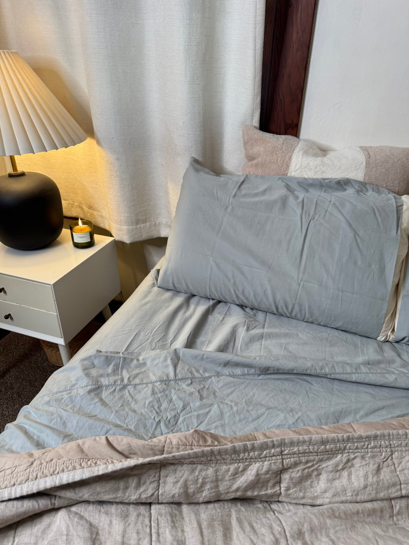 A neatly made bed with light gray sheets, a gray pillow, a black lamp, and a lit candle on a white bedside table.