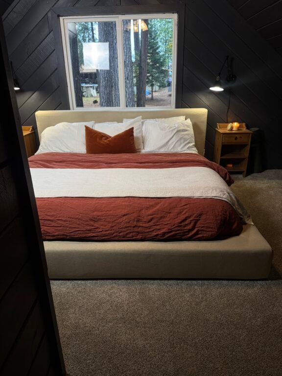 A neatly made bed with white and rust-colored bedding, flanked by two wooden nightstands, sits below a window overlooking trees in a carpeted bedroom.