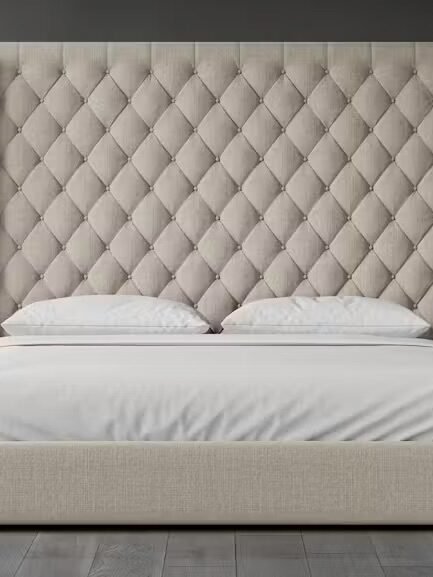 A beige upholstered bed with a tall, diamond-tufted headboard, white pillows, and a white mattress, set against a dark gray wall and wooden floor.
