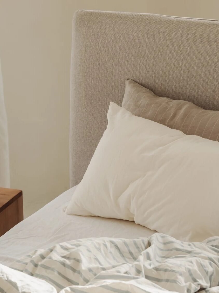Unmade bed with white and striped bedding, two pillows, and a wooden nightstand holding books and a modern lamp in a sunlit room.