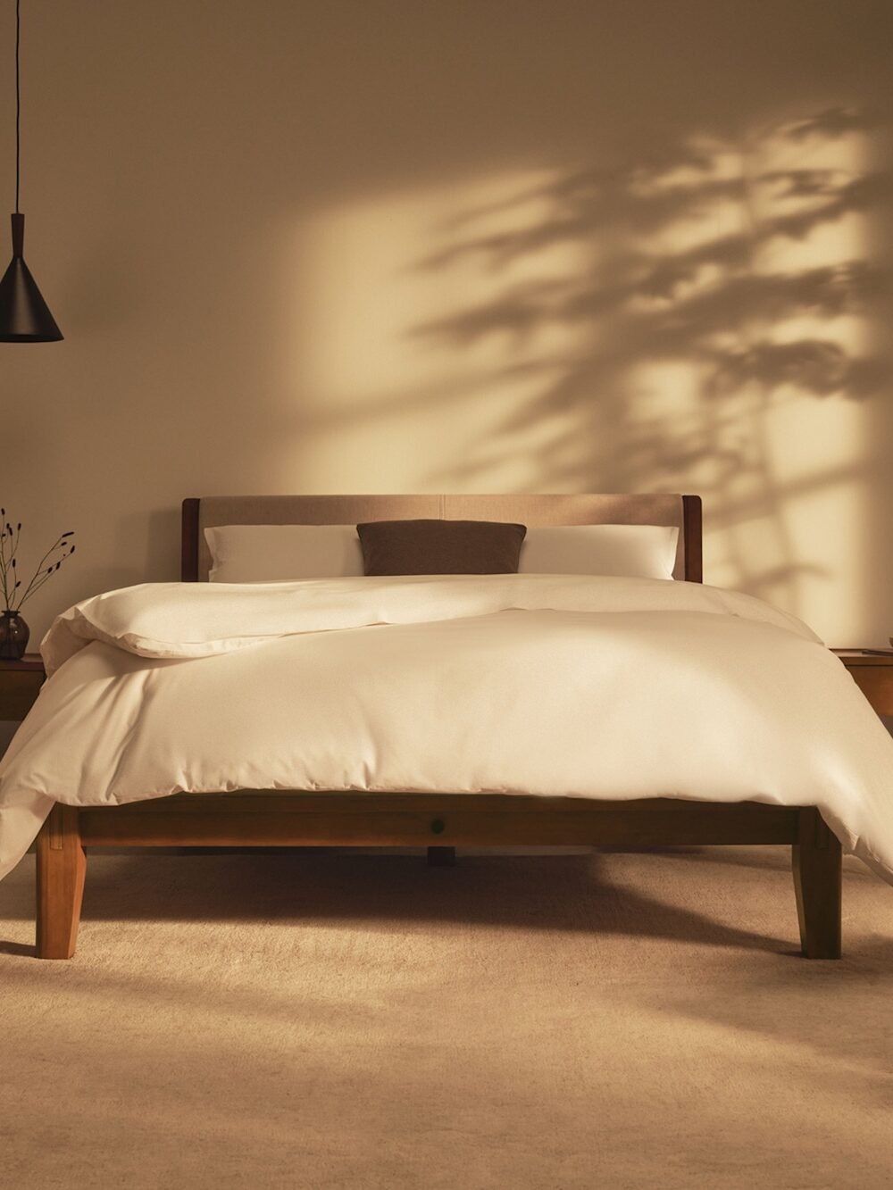 Minimalist bedroom with a wooden bed, white bedding, two nightstands, a pendant lamp, a potted tree, and sunlight casting shadows on the wall.