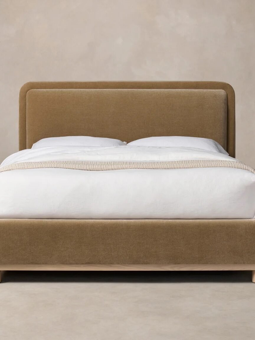 Bed with a tan upholstered frame, white bedding, and light wooden legs, positioned against a beige background.