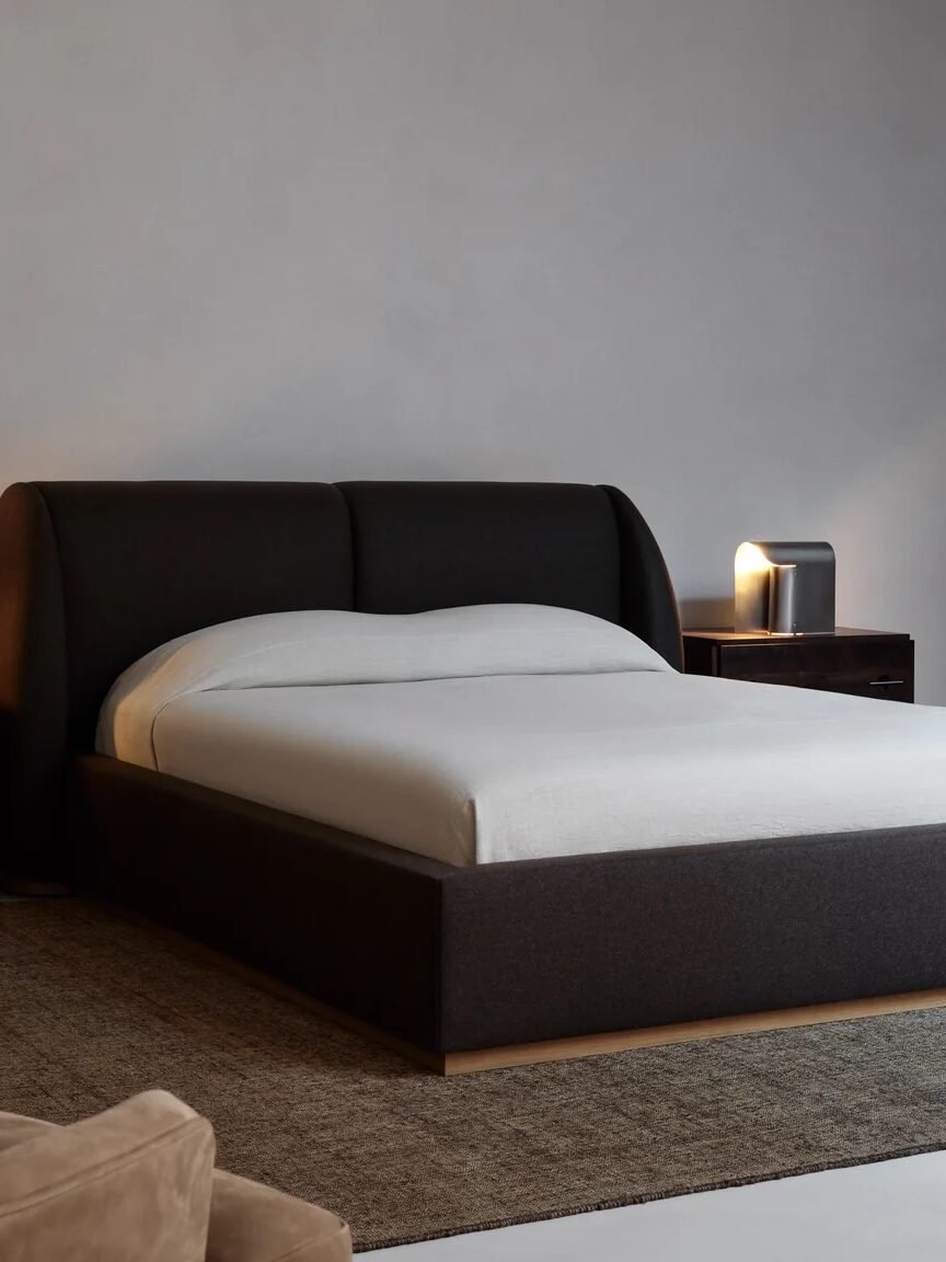 Minimalist bedroom with a dark upholstered bed, two wooden nightstands with lamps, a wooden bench, and a large neutral rug on a concrete floor. Walls are plain and light gray.
