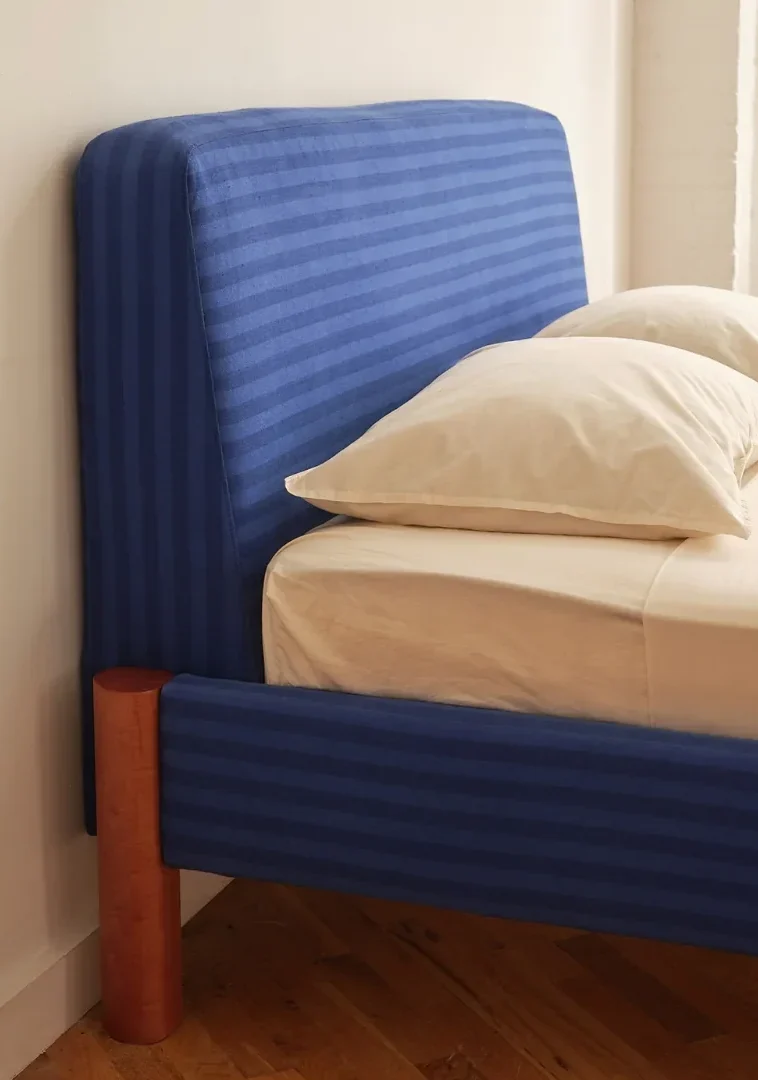 A bed frame upholstered in blue-striped fabric with light-colored wooden legs and cream bedding.