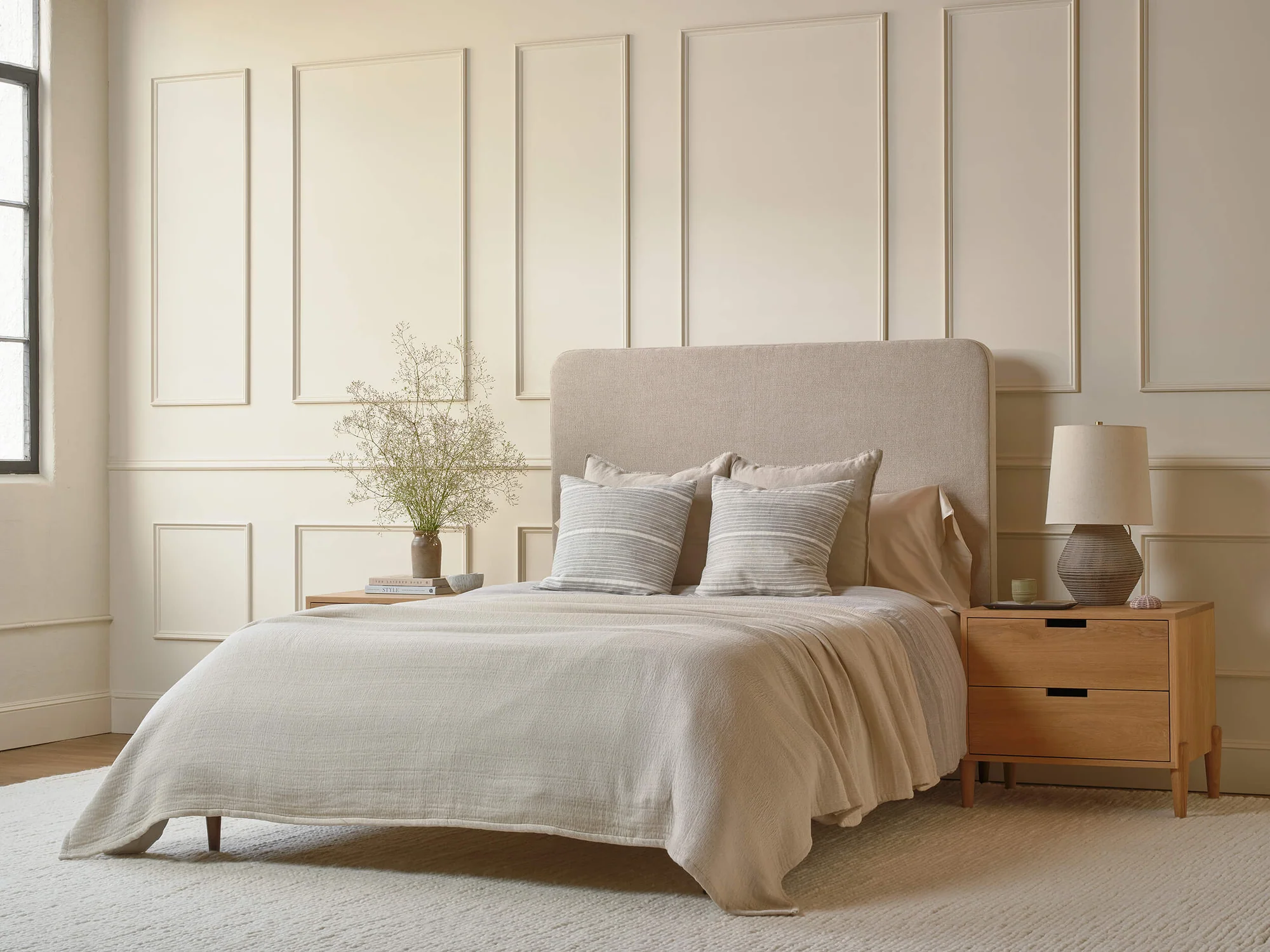 A beige upholstered bed with neutral bedding and striped accent pillows, flanked by two wooden nightstands in a room with paneled walls.