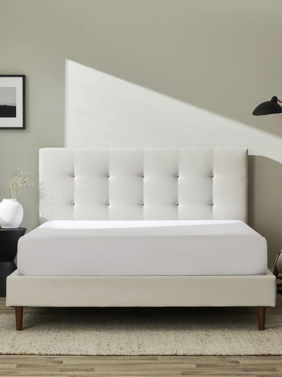 Minimalist bedroom with a beige upholstered bed, tufted headboard, side table with vase, modern lamp, chair, and framed wall art against a light gray wall.