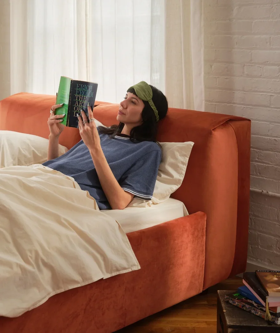A person wearing a sleep mask lies in an orange bed reading a copy of The Art of Loving by Erich Fromm.