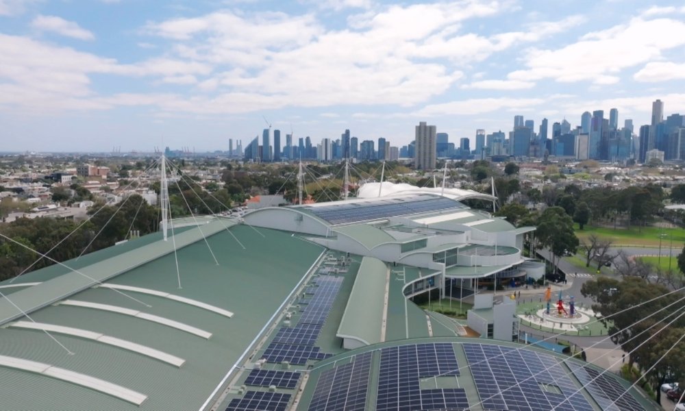 Aerial view of a green-roofed building with solar panels, with a distant city skyline of tall buildings.
