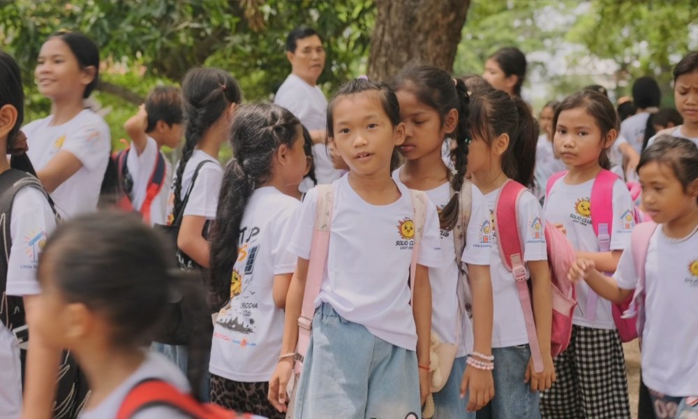 Group of elementary students in white shirts with backpacks outdoors; center girl faces the camera during a school outing.