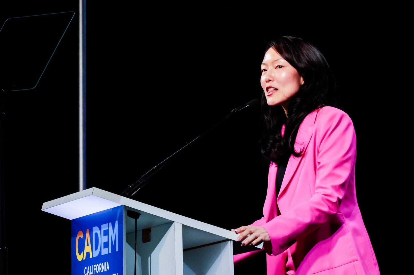 A woman in a pink pantsuit speaks behind a podium that says 'CADEM'