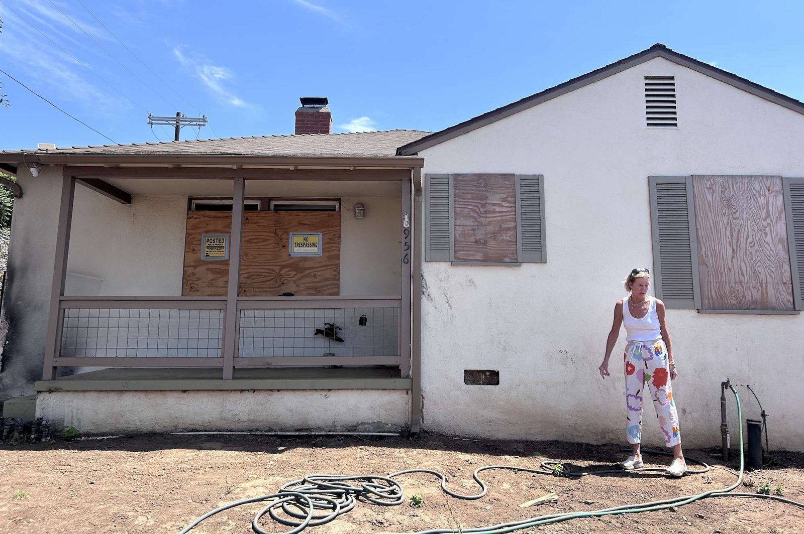 A woman stands in front of a boarded up house near a hose