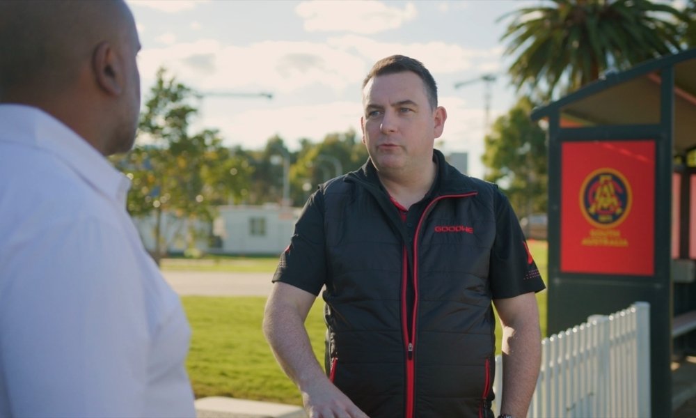 Two men talking outdoors; one facing the camera in a black vest with red zippers, the other in a white shirt.