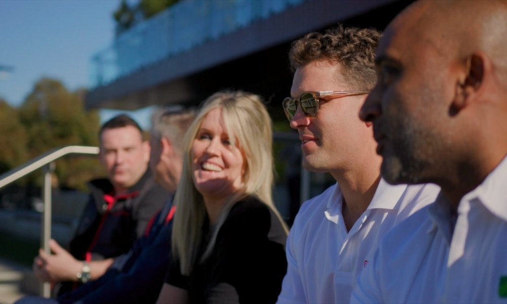Four adults sitting on a bench outdoors, smiling and chatting; focus on a man in a white shirt wearing sunglasses in the foreground.