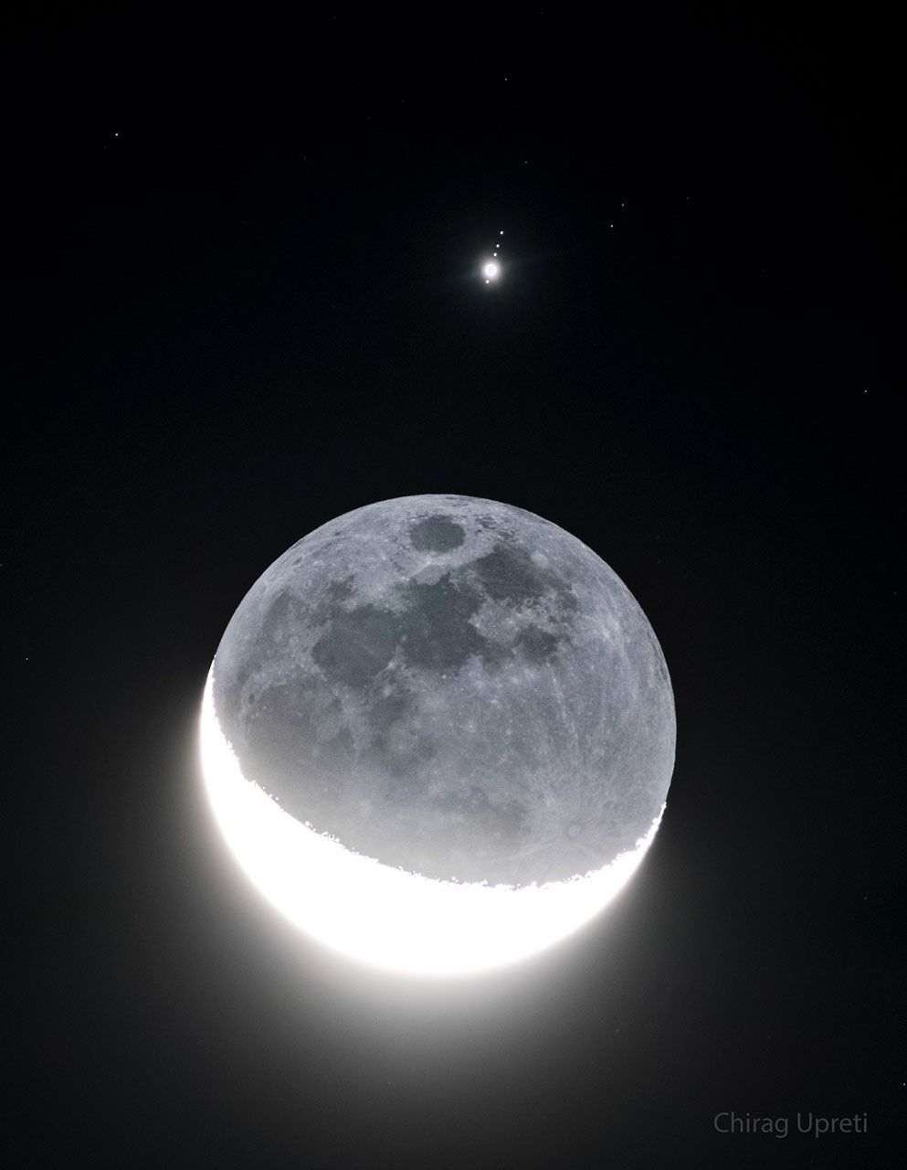 In front of a dark background an image of Earth's Moon appears with a lower crescent brightly lit. The rest of the Moon is also faintly visible. Above the Moon is a bright point of light surrounded by four fainter points of light in row. Please see the explanation for more detailed information.