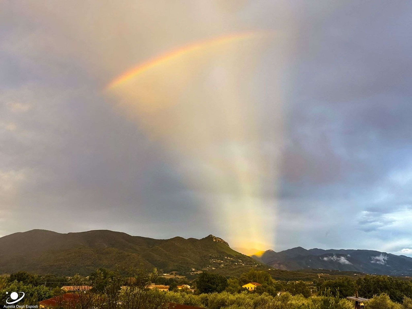 Hills are seen in the distance below a partly cloudy sky. A group of orange rays appears to eminate from one area behind the hills. Across this fan of rays, near the top of the image, a short rainbow can be seen. Please see the explanation for more detailed information.