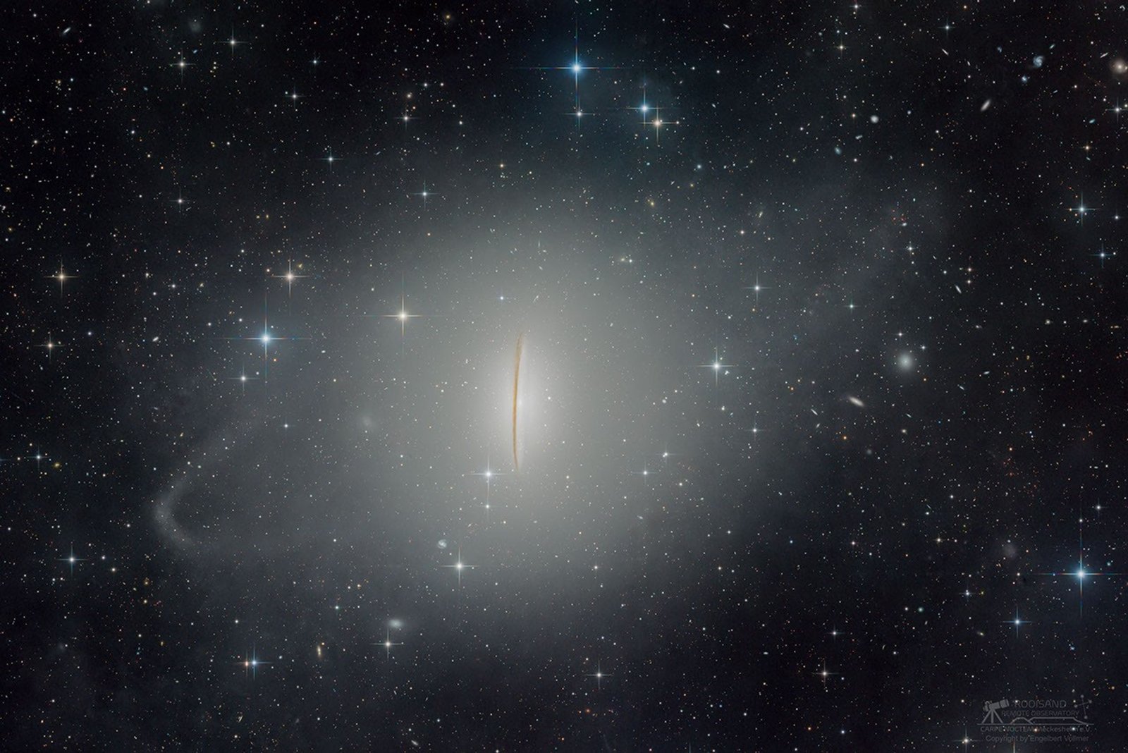 A starfield has an bright blobby mess in middle. Running vertically through the very middle is a dark streak. A diagonal ring of brightness surrounds the central blob.