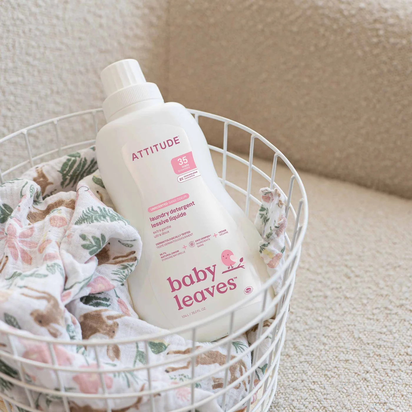 A bottle of Attitude Baby Leaves laundry detergent sits in a white wire basket with a floral-patterned cloth, placed on a textured beige surface.