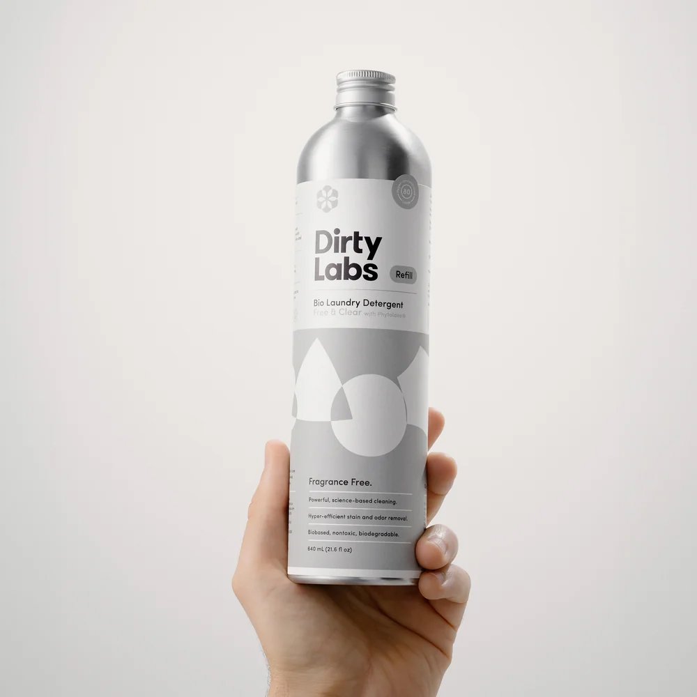 Hand holding a silver bottle labeled “Dirty Labs Bio Laundry Detergent,” fragrance free, with minimalist design against a plain white background.