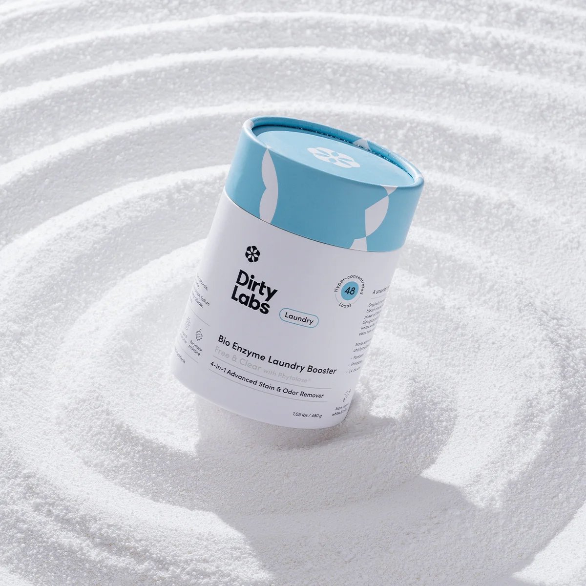 A Dirty Labs Bio Enzyme Laundry Booster container stands upright on a textured, white powdery surface with concentric circular patterns.