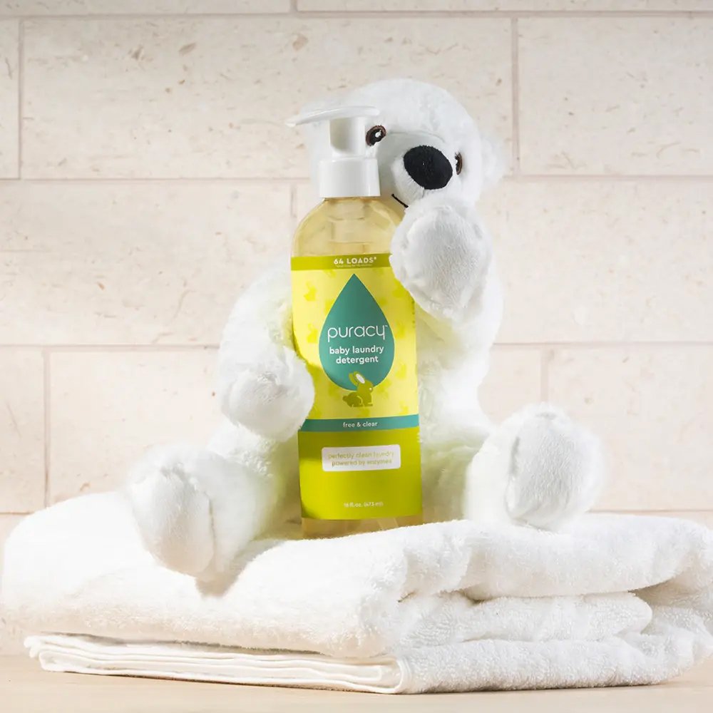 A white stuffed bear sits on folded white towels, holding a bottle of Puracy baby laundry detergent with a yellow and green label against a light tiled background.