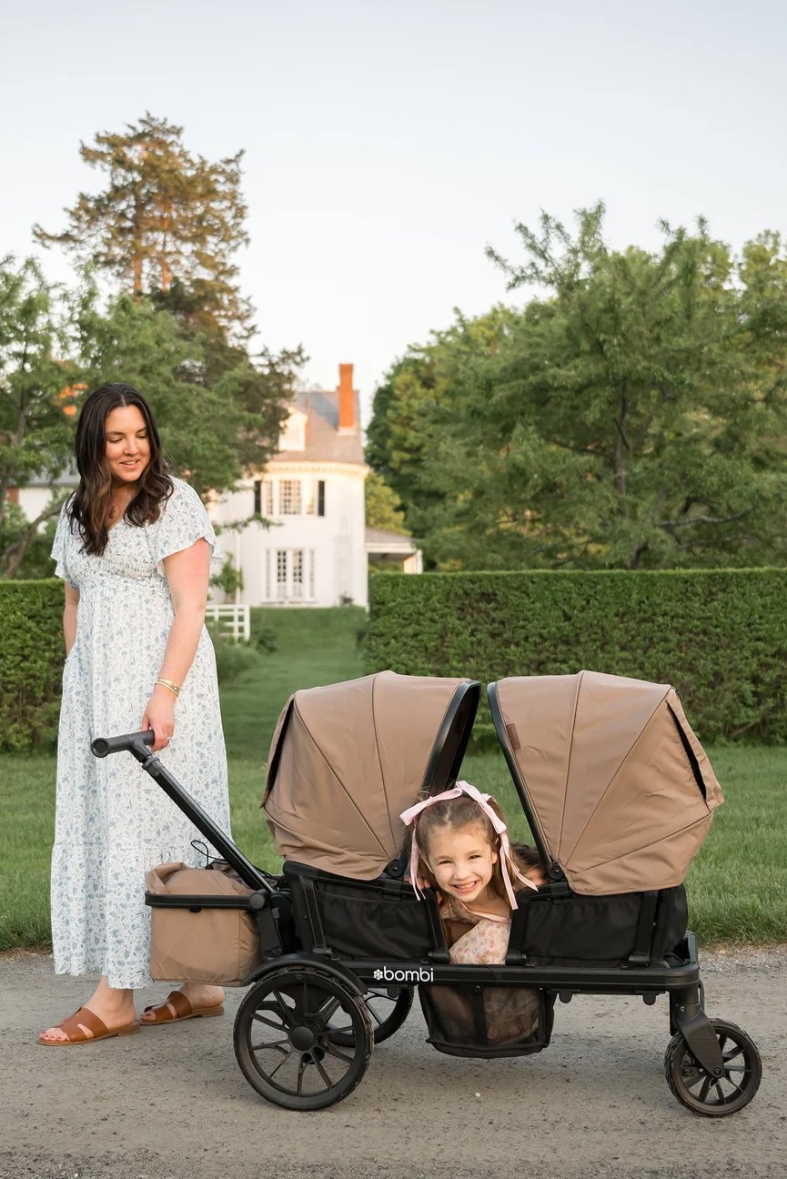 A woman in a light blue dress pushes a tan stroller with two canopies; a smiling young girl peeks out from the front seat. A large white house and green trees are in the background.