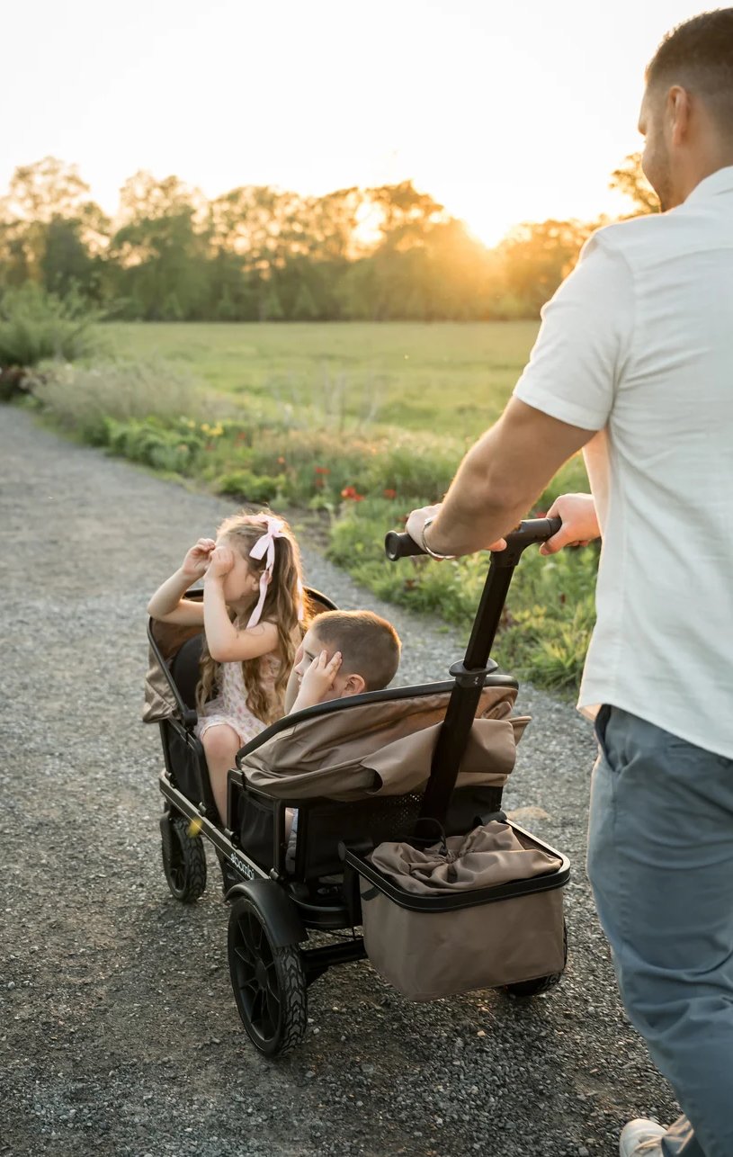 An adult pushes two young children in a brown wagon along a gravel path at sunset, with a field and trees in the background.