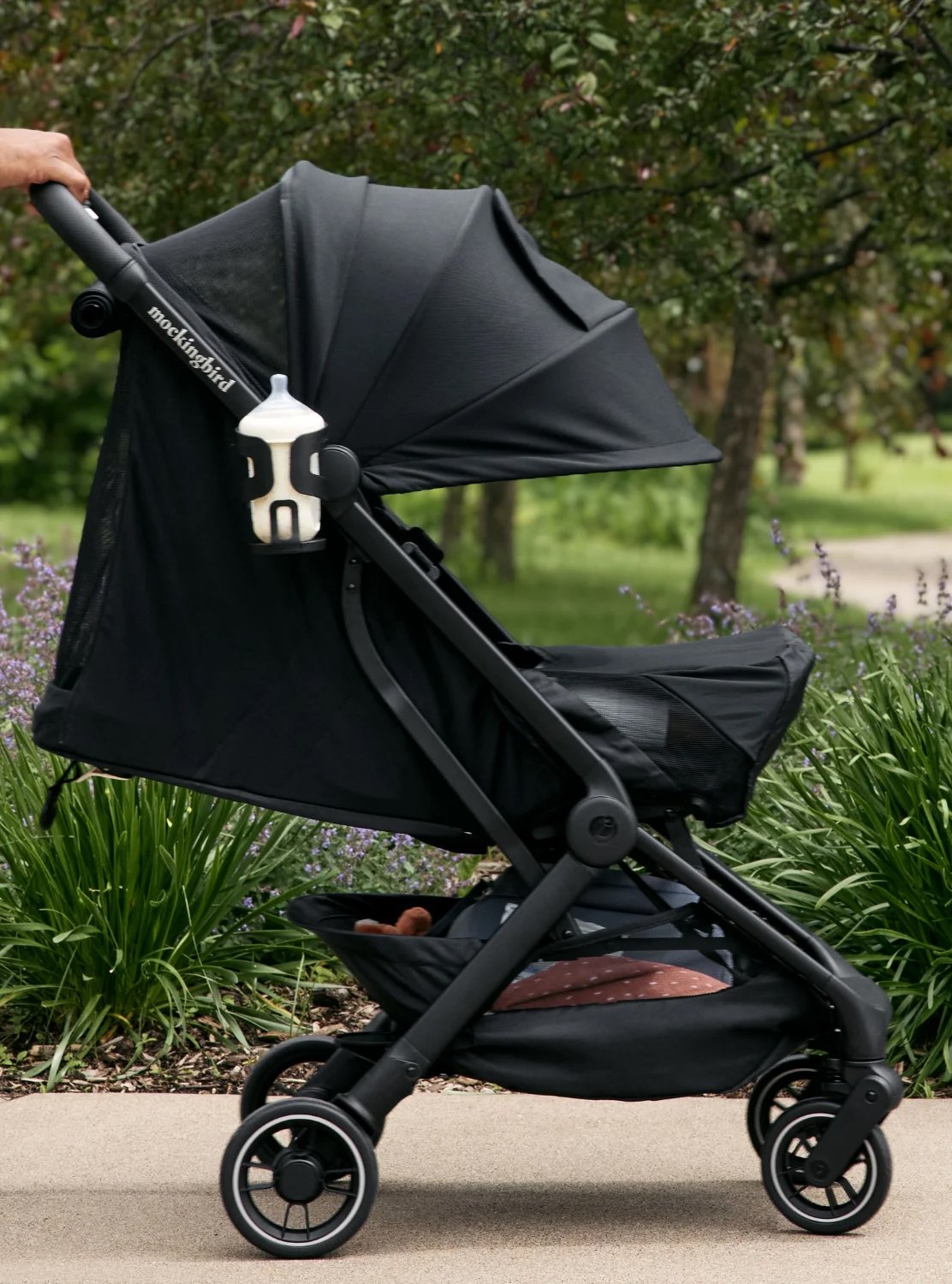 A person pushes a black stroller with a bottle in a holder, along a path bordered by greenery and purple flowers.