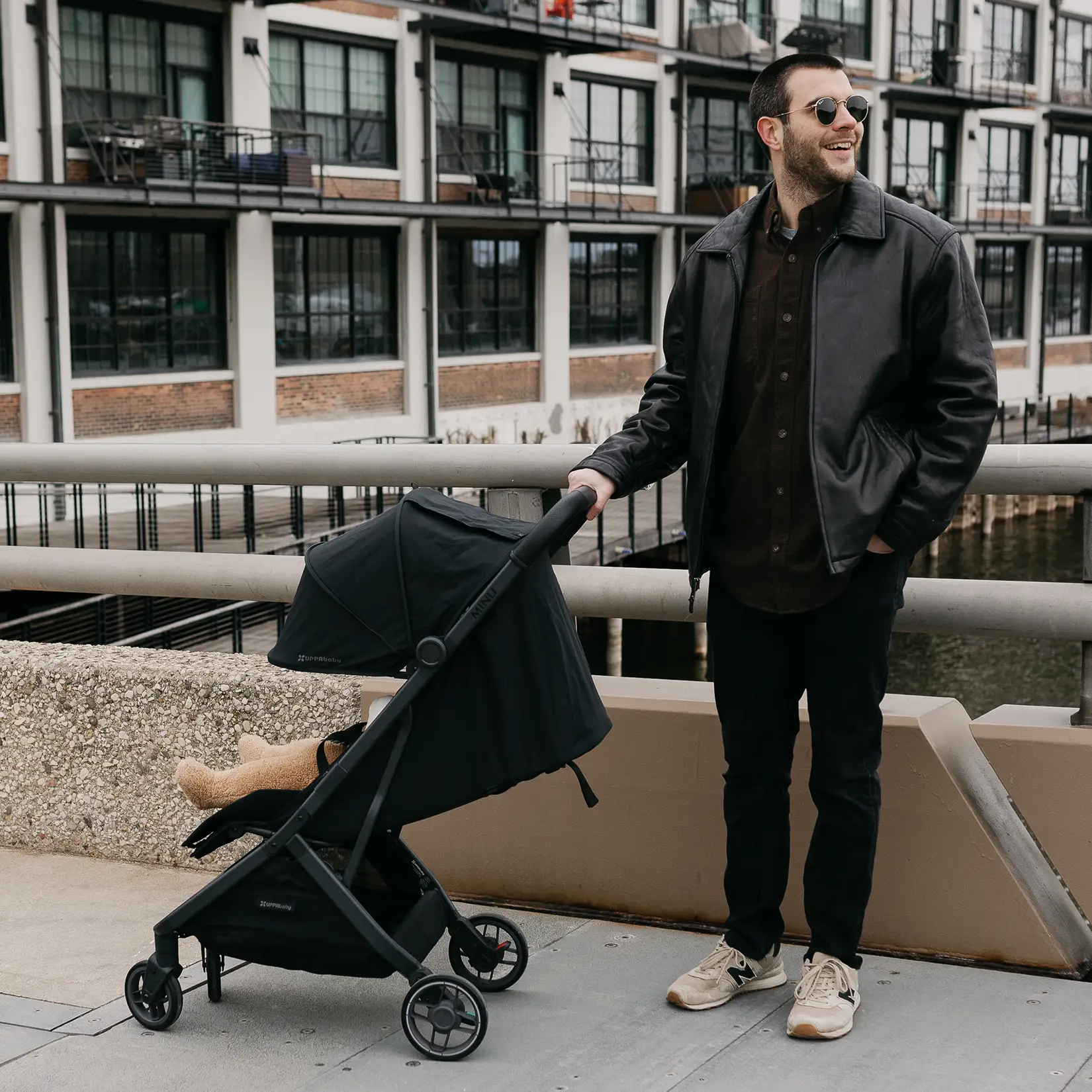 Man in a leather jacket and sunglasses stands on a sidewalk, smiling, while pushing a black stroller with a small child inside near a modern apartment building.