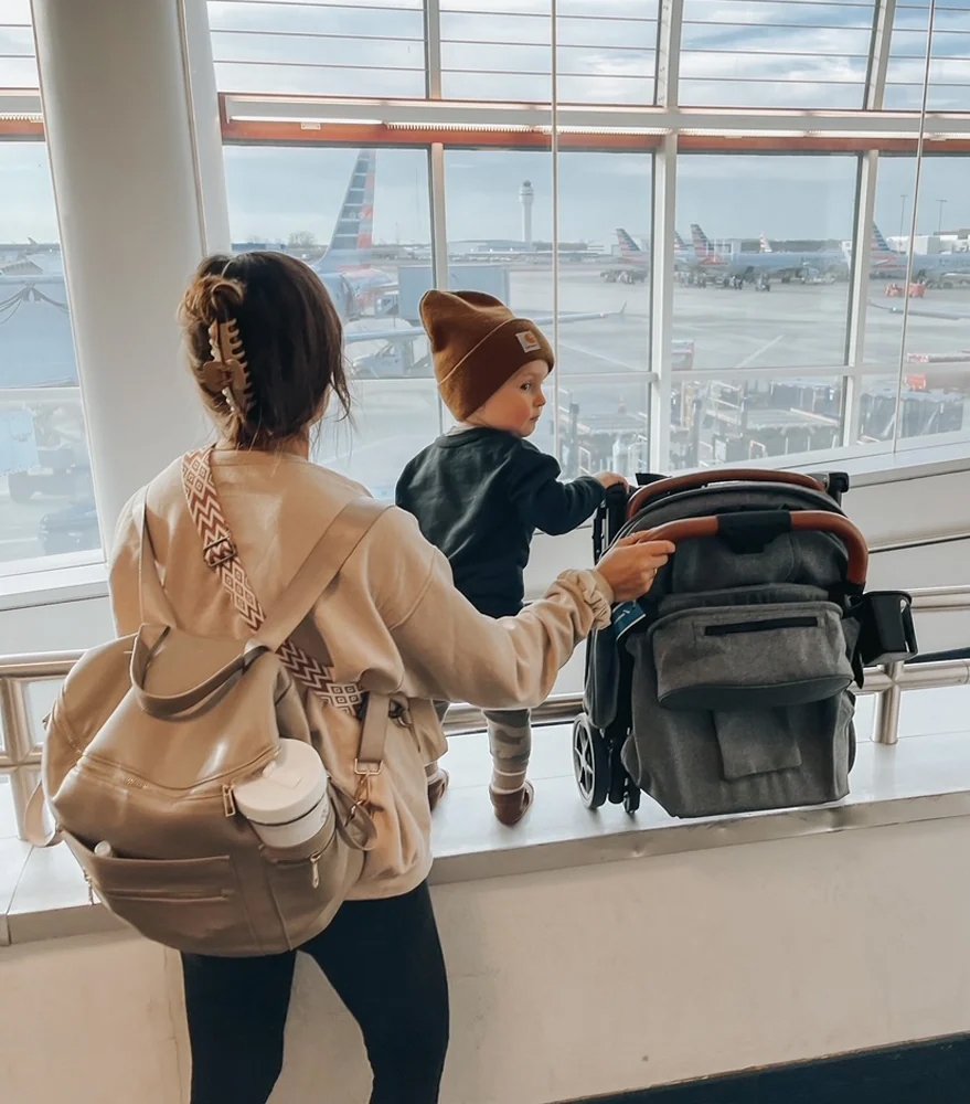 A woman with a backpack stands next to a toddler and a stroller at an airport terminal, looking out at airplanes on the tarmac through large windows.