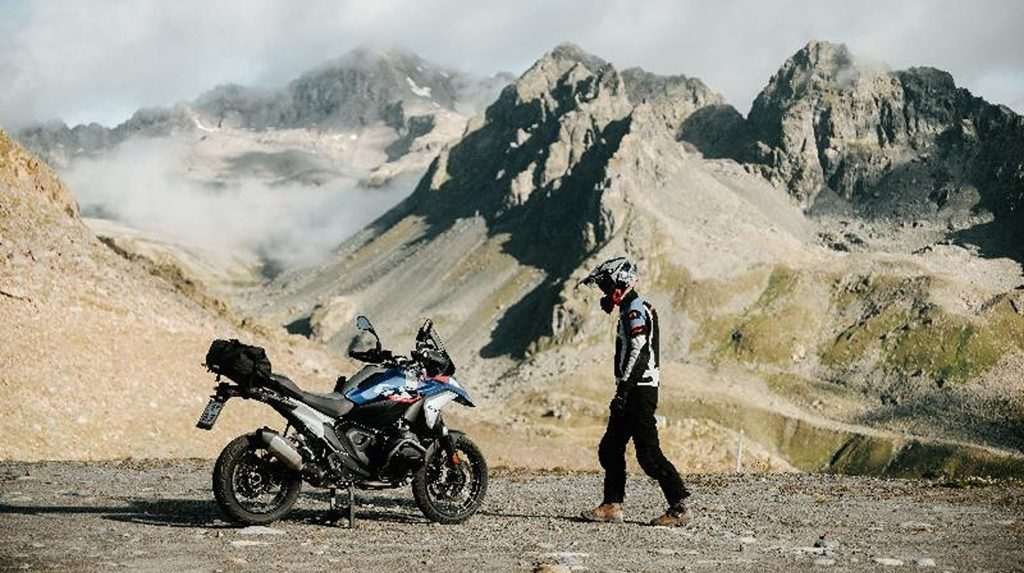 A motorcycle rider with a black helmet on walks towards an adventure motorbike parked on an off-road gravel surface.