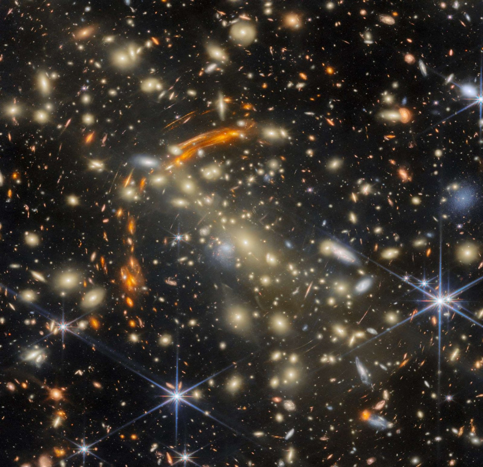 A massive galaxy cluster, made of many golden elliptical galaxies with the largest, brightest one in the center. Heavily distorted orange galaxies appear all around the cluster, the largest concentrated in a large arc around the cluster's center; these are images of very distant galaxies, created by gravitational lensing. A few galaxies are large and bluish-white in color; bright, nearby stars are the same color.
