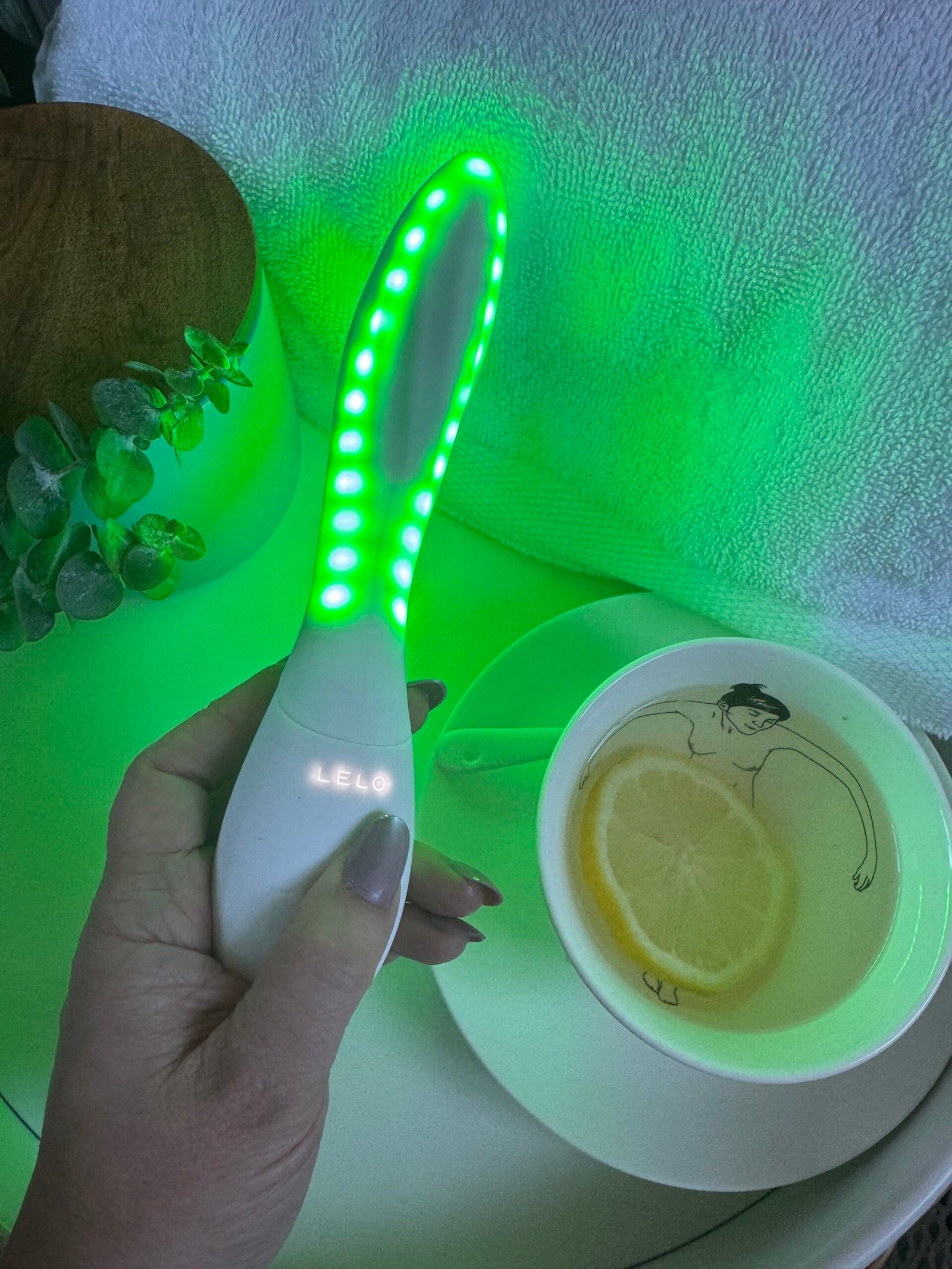 A hand holding a glowing green LELO skincare device next to a teacup with a lemon slice.