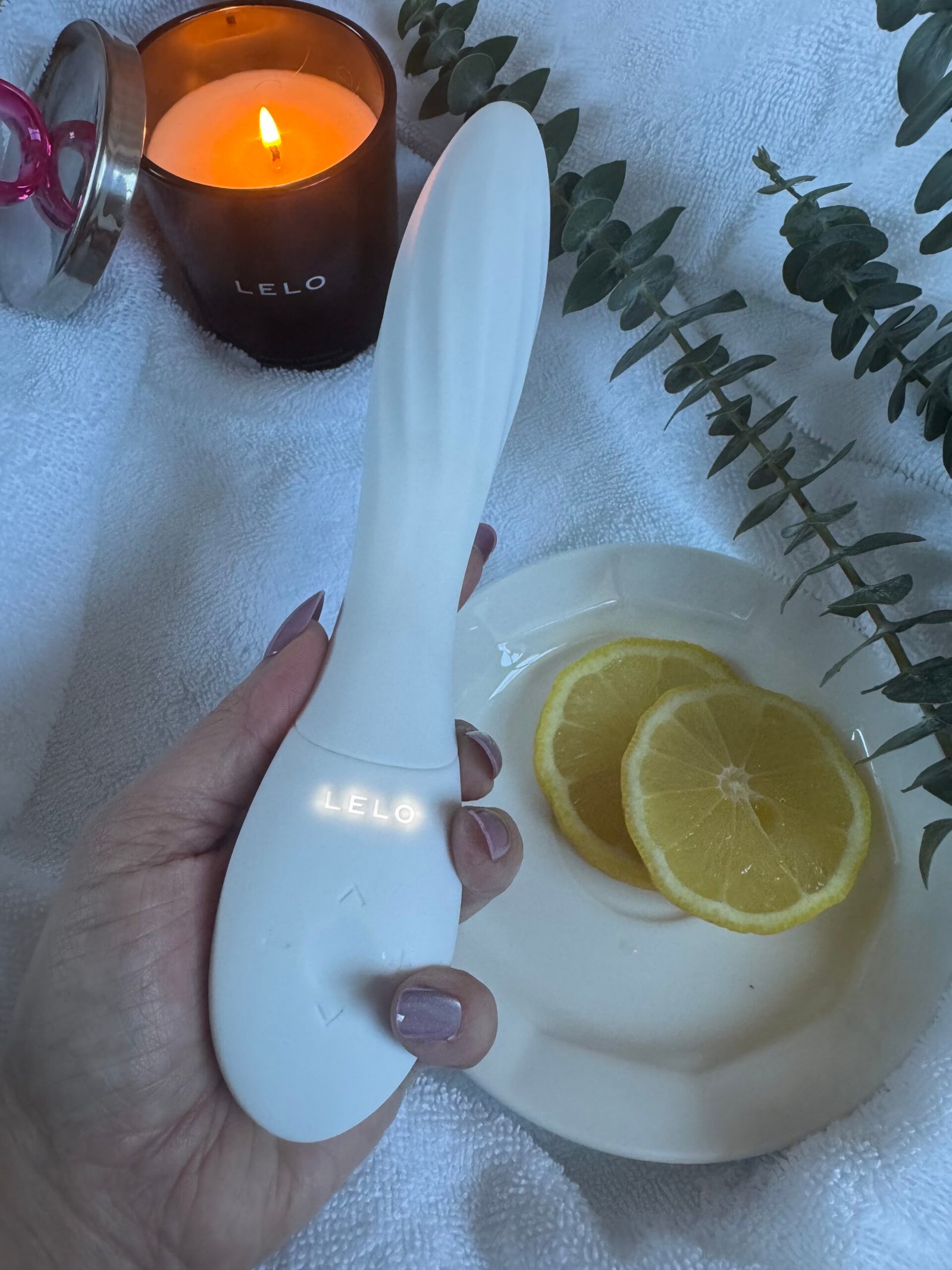 A hand holding a white LELO wellness device, with a lit candle, lemon slices, and eucalyptus sprigs on a white towel.