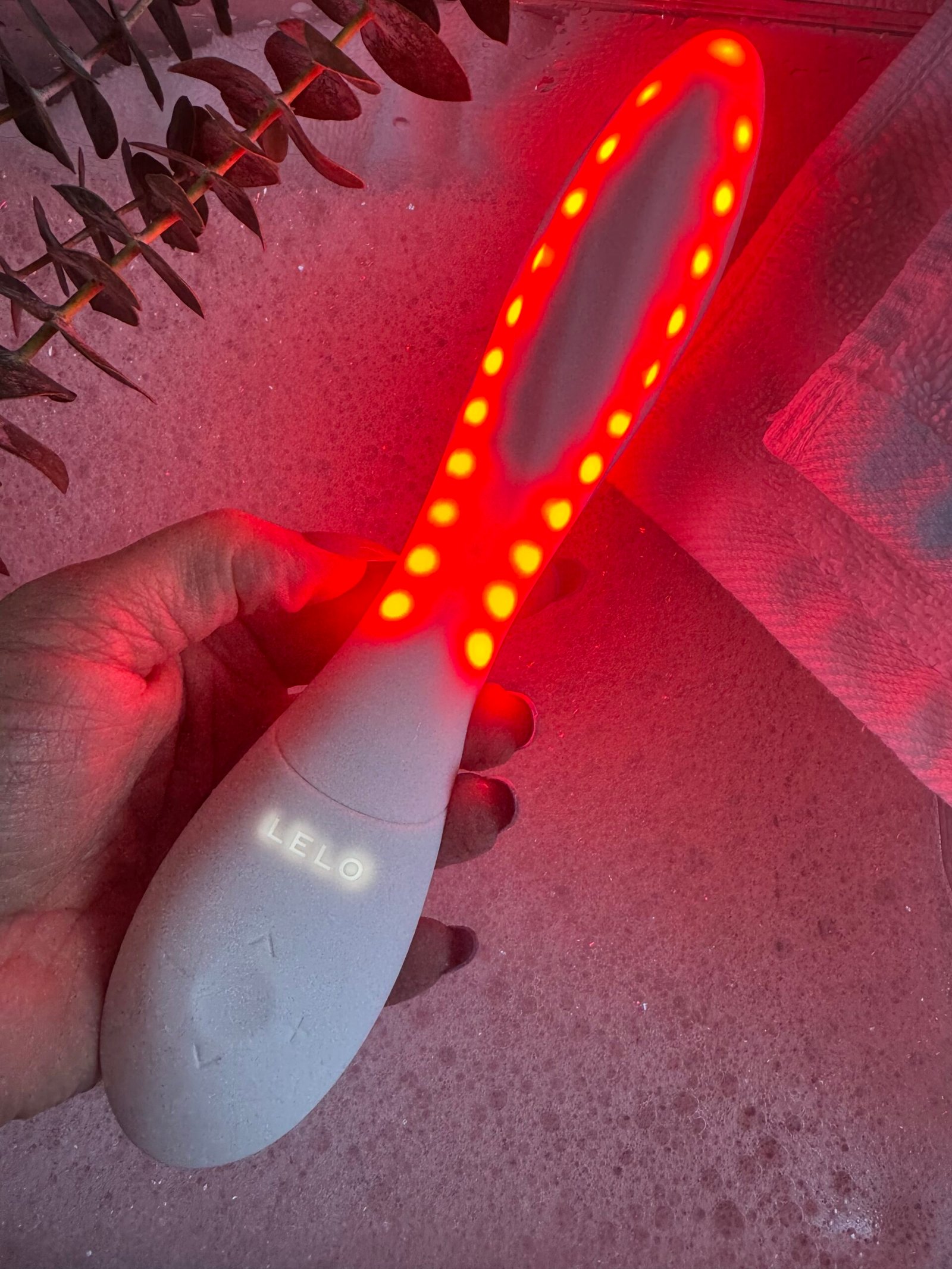 A hand holds a Lelo brand light therapy wand with red LED lights illuminated along its oval head.