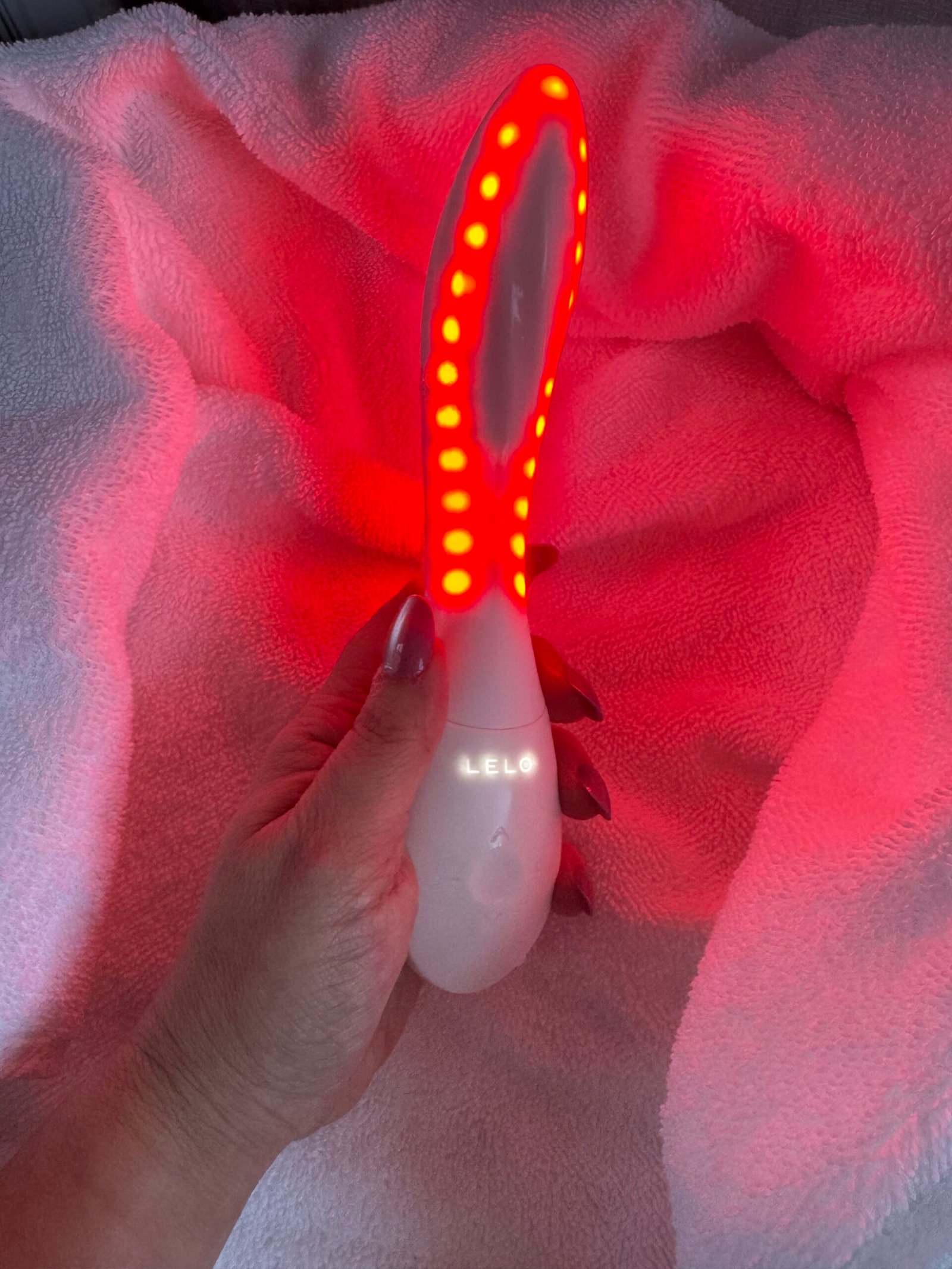 Hand holding a white, curved LELO personal massager emitting a bright red light from a row of LED indicators.